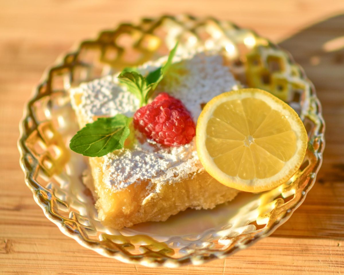 A lemon bar, garnished with basil, a raspberry, and a slice of lemon.